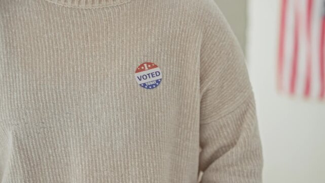 Woman wearing i voted sticker on beige sweater, hand over chest gesture beside a blurred american flag inside a polling building; civic pride.