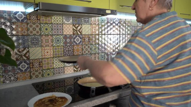 Pour the batter into the pan and flip the pancake. Senior Woman frying thin pancakes in a frying pan. Shrove tide day. Grandmother cook food at kitchen.