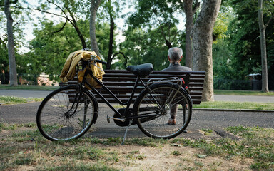 Fototapeta premium Elderly man resting on park bench beside vintage bicycle in urban park