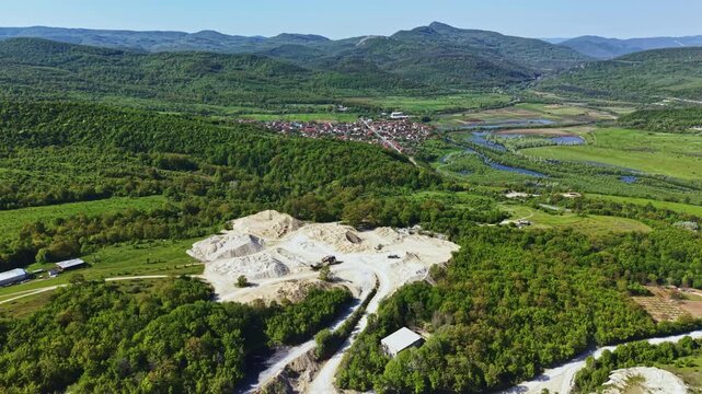 A mining area shows piles of material on land. The site is set in a valley with green hills on all sides. A small town and bodies of water are visible in the distance.