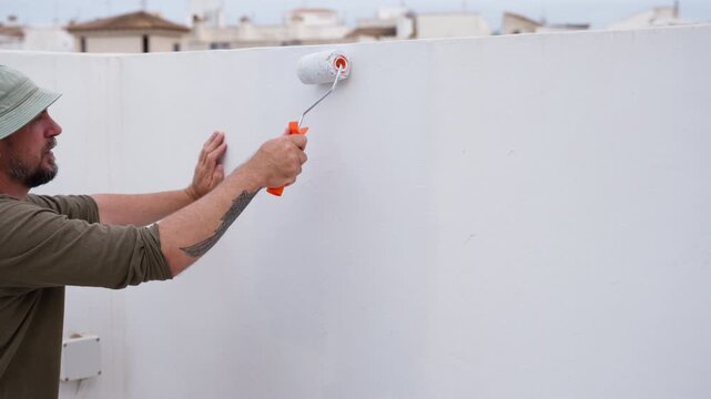 Man using paint roller to paint rooftop terrace wall during DIY renovation project. Home repair, maintenance work, and residential improvement concept.