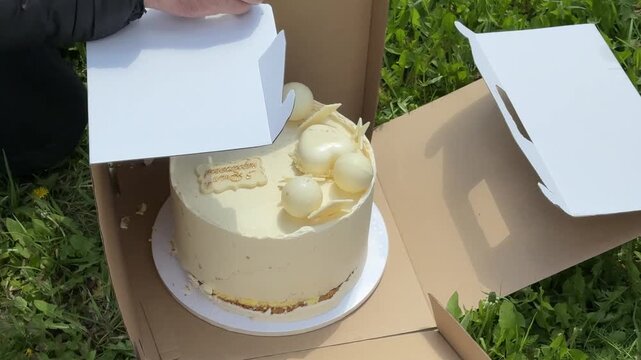 Male hands fixing a damaged birthday cake box after a white 70th birthday cake fell on grass outdoors