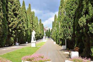 Fototapeta premium Beautiful landscaped alley lined with tall evergreen cypress trees and classical statues, peaceful park walkway with benches and flowers on a sunny day with storm clouds