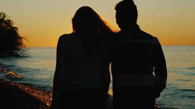 Silhouette of couple walking along pebbled beach at sunset holding hands with bright sun reflection shimmering on water waves during golden hour summer evening 