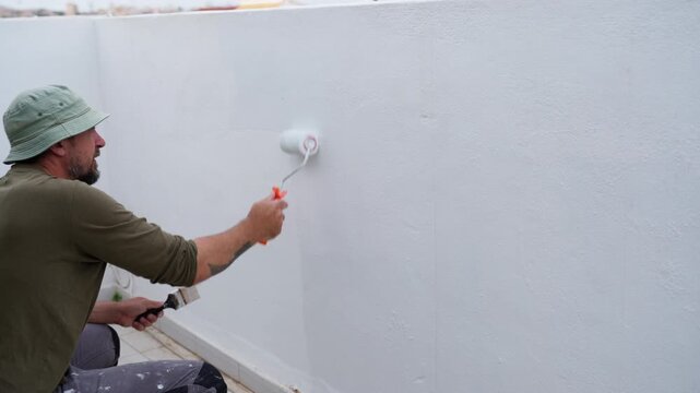 Construction worker performs exterior renovation by applying protective coating to rooftop terrace wall using roller. Building maintenance, repair, and home improvement concept.