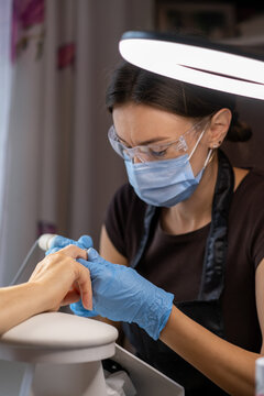 Female nail technician applies manicure treatment to client's hand under a bright lamp in a beauty salon, showcasing modern tools and equipment