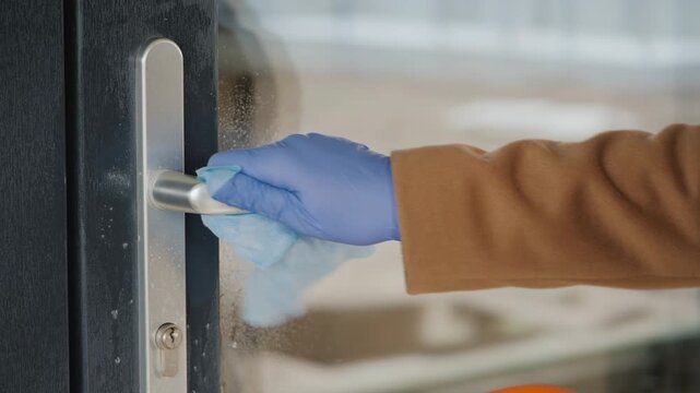 Close up of person wearing blue gloves spraying disinfectant onto door handle and wiping it clean to prevent germ spread during pandemic.