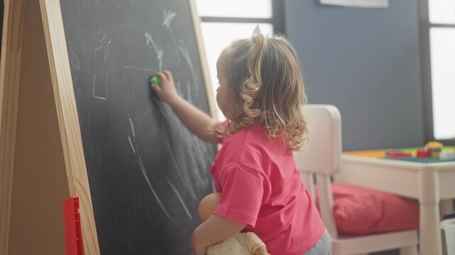 Girl erasing chalkboard with eraser while clutching doll inside a childcare building; curiosity focused learning.