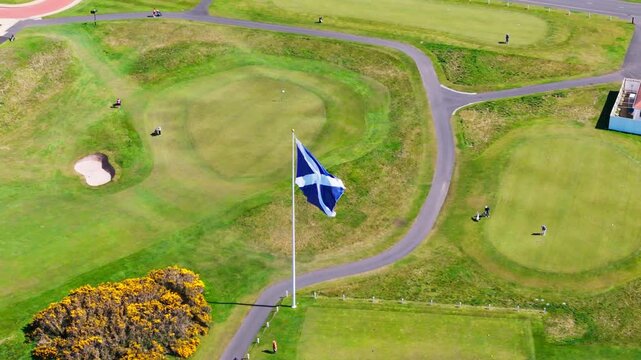 Slow motion aerial shot of the blue and Saltire flag waving in the wind above the lush fairways and greens of a coastal golf course in Scotland.