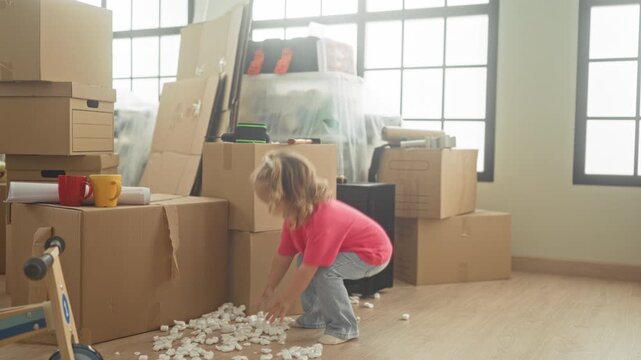 Little girl picking up packing peanuts with bare hands amid stacked moving boxes and a wooden scooter in a building living room; playful curiosity.