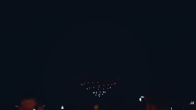 illuminated aerial drone show at glowing night sky, programmed flying robotics light performance over city, people watching and filming, advanced technology concept