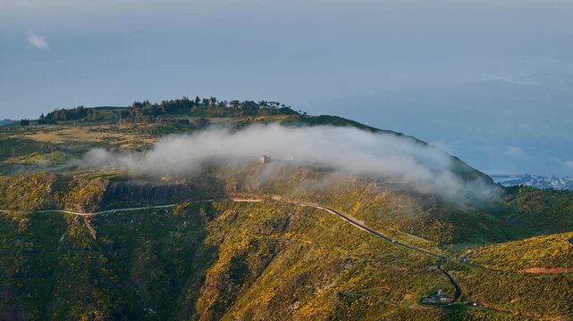 Winding mountain road leading to Pico do Arieiro, Madeira Island, with low cloud drifting across green volcanic slopes, aerial drone view