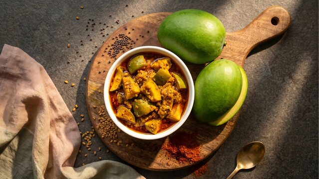 Overhead view of a white bowl filled with indian mango pickle, surrounded by whole green mangoes, scattered spices, and a textured cloth on a wooden board