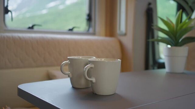 Hand places two mugs on a table and pours coffee inside a bright camper kitchen. Cozy off-grid van life scene with warm, minimal interior and relaxed morning routine vibe.