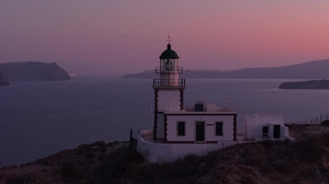 Drone orbiting the Santorini lighthouse at sunrise with a romantic pink horizon, capturing the caldera cliffs, Aegean Sea, and dreamy island landscape.