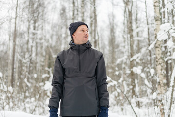 Man in a membrane sportswear jacket standing in a snowy winter forest. Male wearing technical gear with a high collar amid snow covered trees. Confident hiker showcasing functional apparel on cold day © Sk Elena
