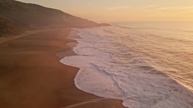 Drone pullback over Praia do Salgado in Nazare, Portugal. Cinematic aerial view of waves crashing on the sandy shore during golden hour sunrise