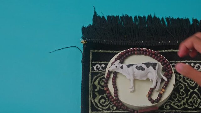 Hand places spotted cow figurine on pedestal by prayer beads and prayer rug on blue background