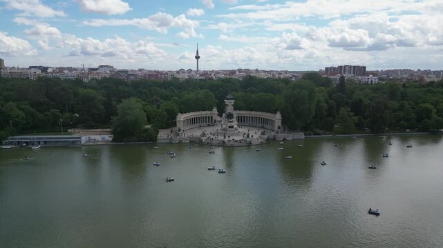 The Alfonso XII monument with boats on the lake at El Retiro Park. Cloud-filled sky and city skyline in the distance. Suitable for travel and cultural content.