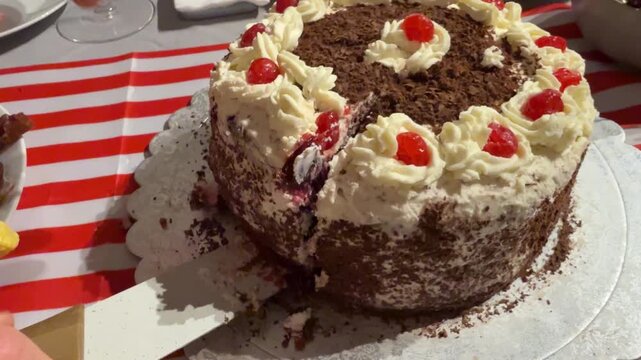 Chocolate forest cake on white doily, whipped cream rosettes with cherries and chocolate shavings, hands reaching to serve dessert at party table
