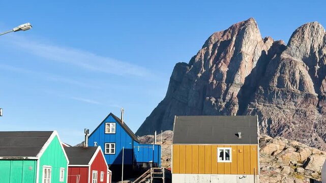 Colorful houses sit on the rocks near the ocean in Greenland. Icebergs float in the water. The sun shines brightly. A few boats are seen near the shore. This location shows daily life by the sea.