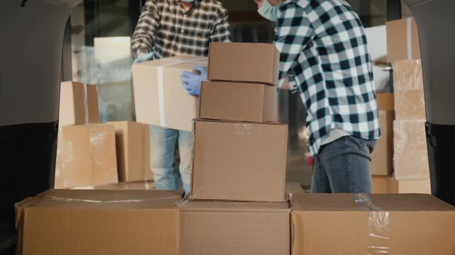 Two male warehouse workers wearing masks and gloves loading cardboard boxes into a delivery van for shipment.