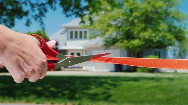 Person cutting red ribbon with scissors in front of new residential house during grand opening ceremony footage.
