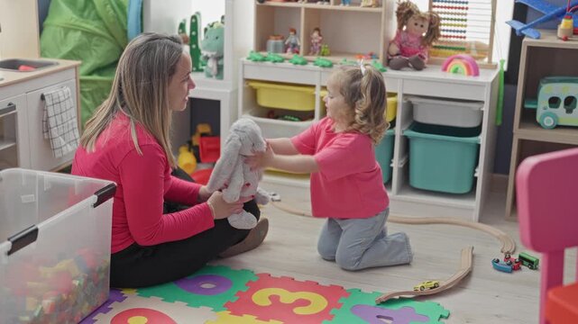 Woman sitting on floor passing a gray stuffed rabbit to little girl, hand extended near toy shelves in playroom building; nurturing bond.