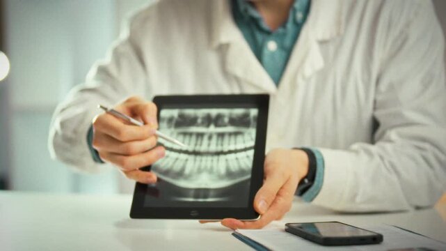 Examining patient's X-ray, doctor in crisp white coat sits at a table in a bright medical office. Using a tablet, he highlights problem areas and discusses tailored treatment options with the patient