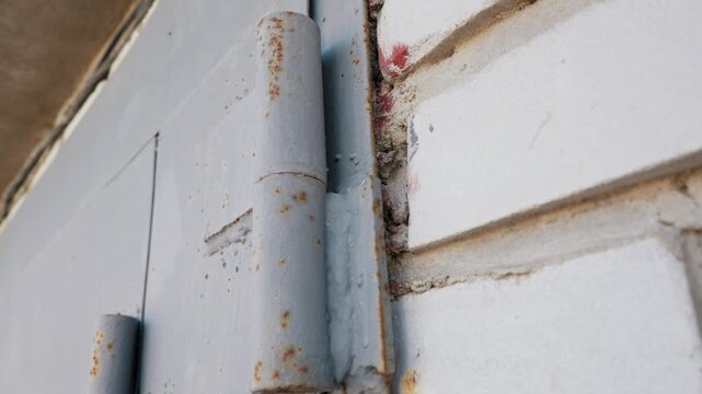 A close-up view shows a rusty metal hinge securing a heavy grey painted door to an old white brick wall. Concept of urban decay, aging infrastructure, and reliable physical security.