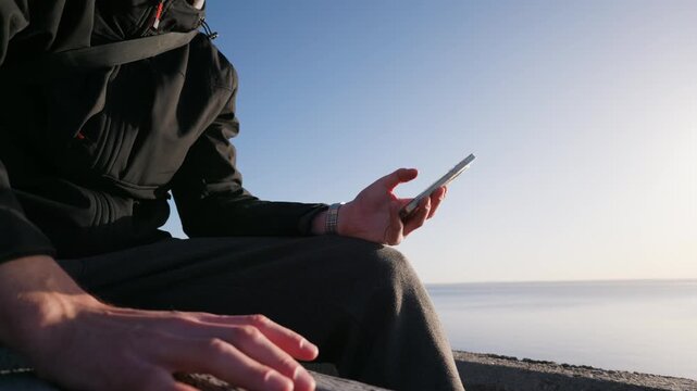 An unrecognizable man sits outdoors by the water and scrolls on his smartphone. He taps his fingers rhythmically on his leg under a clear blue sky.