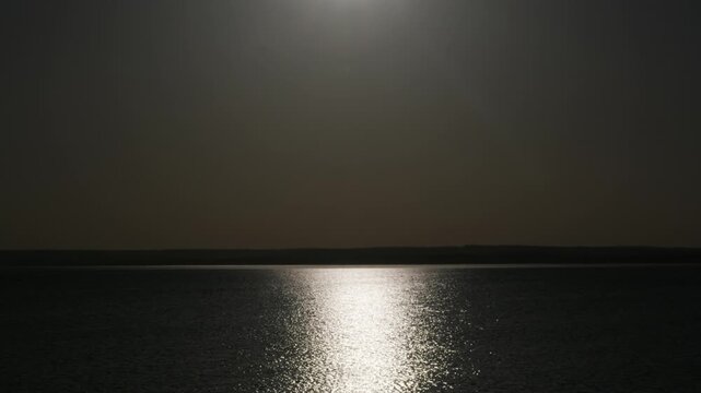 Dark ocean surface shimmers gently under a bright light source, creating striking high contrast reflections across calm waves. Concept of tranquil solitude and deep atmospheric mystery.