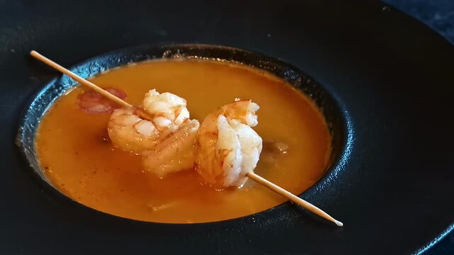 A rich, orange seafood soup served with a wooden skewer of two grilled shrimps.