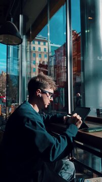 Young male creative professional with wireless earbuds drawing on a digital tablet with a stylus, sitting by a large window with city street reflections in a modern urban coffee shop
