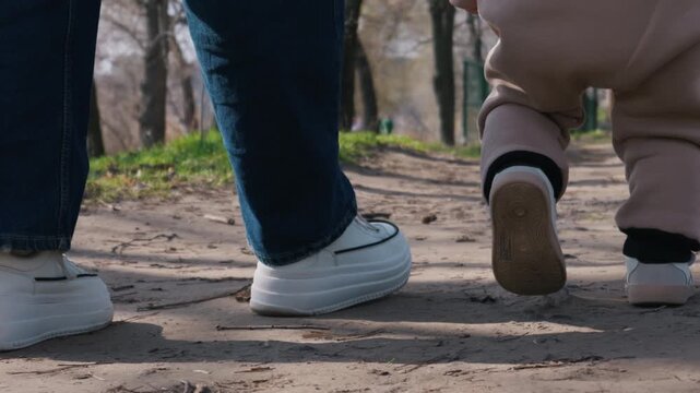 A low angle view captures a toddler in beige sweatpants taking careful steps along a sunlit dirt path. An adult in blue jeans and white sneakers follows closely behind for support.