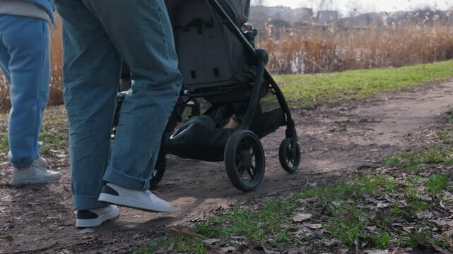 Low angle tracking shot of parents legs walking with baby strollers along a dirt path near a pond. This portrays the concept of active parenting and enjoying family leisure time outdoors.