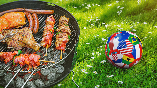 Delicious grilled meat with vegetables sizzling over the coals on barbecue with a soccer covered with multiple national flags symbolizing an international football tournament.