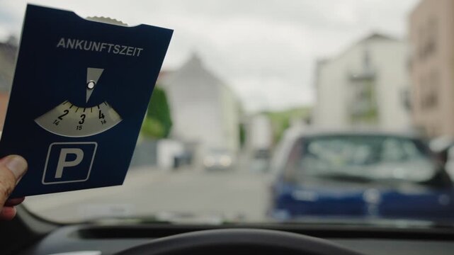 Driver holding German parking disc with word Ankunftszeit meaning arrival time inside car before parking in European city street