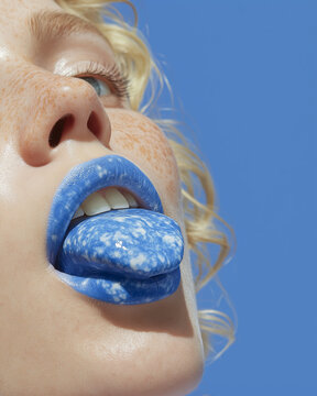 Macro beauty image of glossy blue lips holding a speckled blue tongue against a blue backdrop. Striking fashion visual for cosmetics ads and editorial design.
