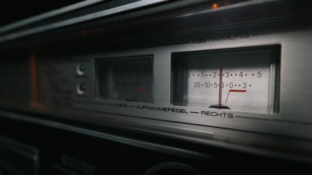 Illuminated analog recording level meter on a silver vintage stereo deck. The glowing red needle rests on the dial in the dark. Represents a retro analog audio concept.