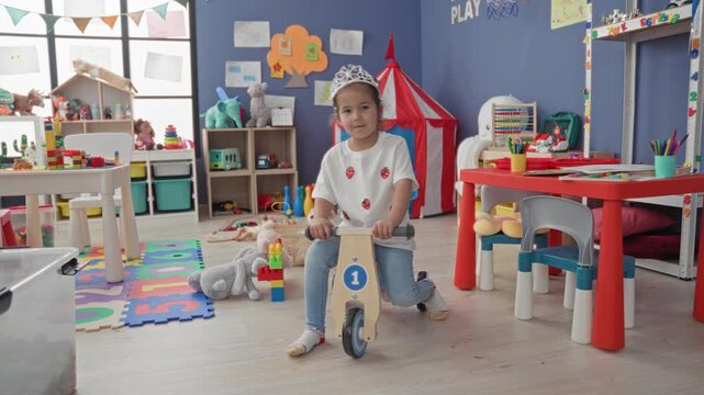 Girl on a wooden balance bike holding handlebars in a colorful playroom building with toys, table, and tent visible; playfulness imagination.