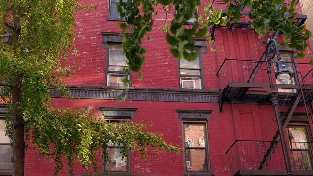 Historic red brick apartment building with fire escape and green trees on sunny street, classic New York City residential architecture facade