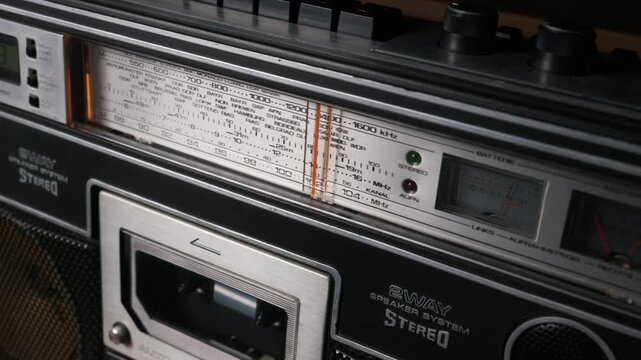 A close-up shot of a vintage analog radio tuner display on a boombox. The brushed metal faceplate lists various European stations and frequency bands. The retro device evokes a sense of nostalgia