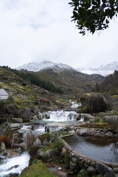 View of Praia Fluvial de Loriga with cascading waterfalls, stone pools, and large boulders against a backdrop of snow-capped mountains in Loriga, Distrito da Guarda, Portugal.