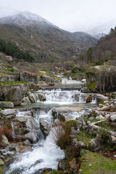 View of Praia Fluvial de Loriga with cascading waterfalls, stone walls, and snow-capped mountains under a misty sky Loriga, Distrito da Guarda, Portugal.