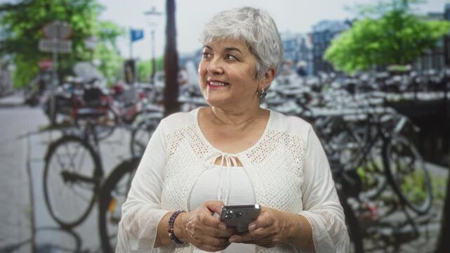 Woman with grey hair holding smartphone in both hands on street, smiling while texting; contentment connection.