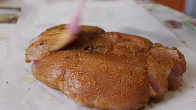 Close up view of a hand using a pink silicone pastry brush to evenly spread a spice marinade over a large piece of raw meat on parchment paper. This depicts a food preparation concept.