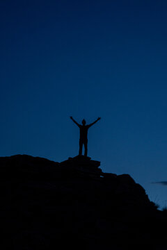 View of the silhouette of a man standing on a rocky ridge with arms outstretched against a dark blue twilight sky in Moab, Utah, United States.