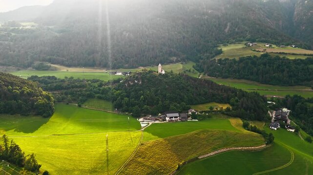 Hilltop chapel in South Tyrol, Italy with sunlight breaking through clouds over rural Dolomites landscape, aerial view