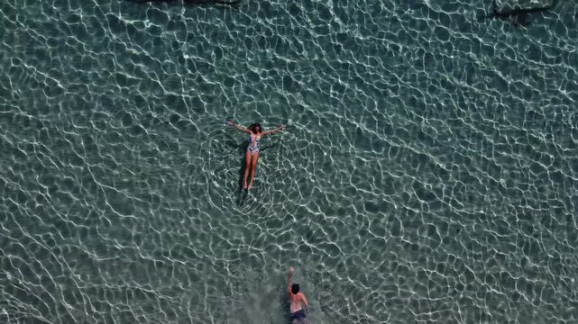 Woman floating alongside a man swimming in turquoise sea water on vacation, Greek Island, Paros, Overhead aerial